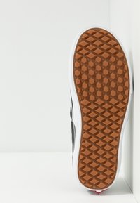 Podeszwa czarnych sneakersów z białą środkową częścią, brązowy bieżnik z wzorami w diamenty i sześciokąty, stojąca pionowo w kącie.