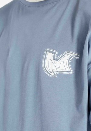 Lichtblauw T-shirt met een wit en grijs abstract geborduurd patch op de linkerborst, met zichtbare stikdetails en een zachte stoftextuur.