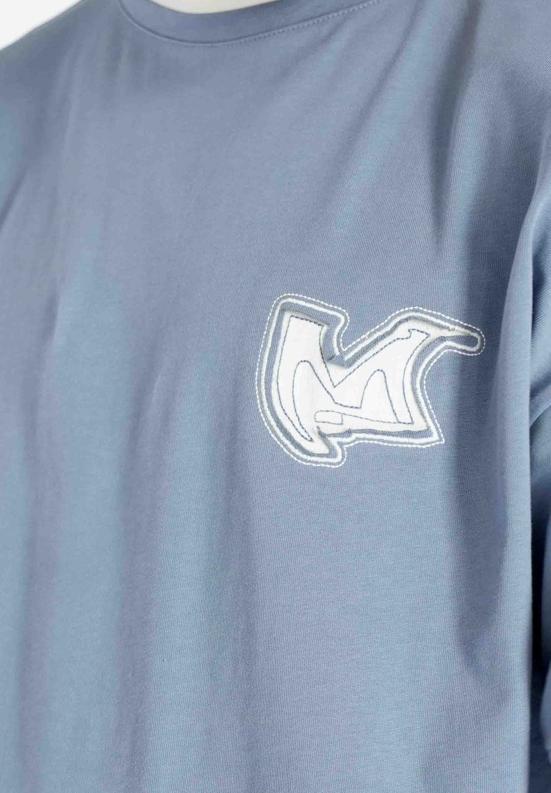 T-shirt bleu clair avec un patch brodé abstrait blanc et gris sur la poitrine gauche, montrant des détails cousus et une texture de tissu douce.