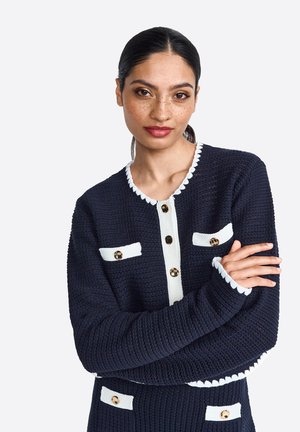 Navy knitted cardigan with white accents, featuring gold buttons and pockets, textured fabric, and scalloped detailing on the collar and cuffs.