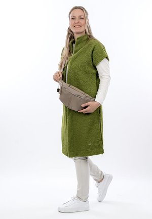 Kvinde iført en grøn tekstureret ærmeløs frakke over hvide lange ærmer og bukser, der holder en beige tværgående taske og står på ét ben.