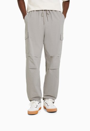 Mann trägt hellgraue Cargohosen mit Seitentaschen und weiße Sneakers mit Gummisohlen, Hände in den Taschen, vor einem schlichten weißen Hintergrund.