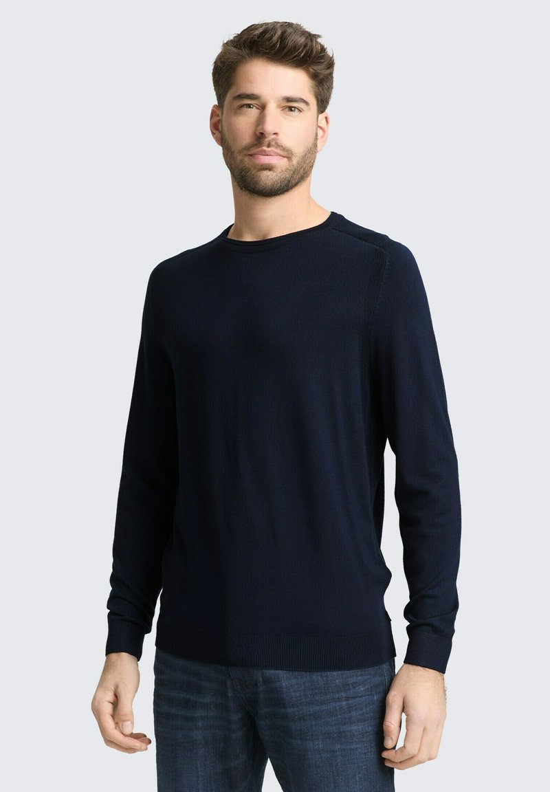 Mężczyzna w ciemnoniebieskim swetrze z długim rękawem i niebieskich dżinsach, stojący na tle jasnoszarego tła, zwrócony przodem z neutralnym wyrazem twarzy.