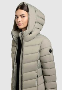 Svetlo siva puffer jakna z velikim kapuco, horizontalno šivenje, sprednji zadrgo in stranski žep. Ima črne kovinske dodatke in podrobnosti slog. 