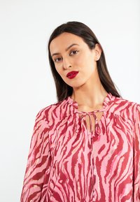 Blusa rosa con un motivo a strisce di tigre in rosa scuro e riflessi dorati. Presenta una consistenza morbida, un tessuto leggero e un dettaglio con fiocco al collo.