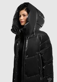 Chaqueta de plumas negra con capucha grande, acabado brillante, bolsillos con cremallera y diseño acolchado. Presenta una etiqueta de marca prominente en el costado.