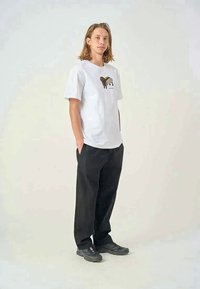 T-shirt branca de manga curta com um gráfico colorido de um cão, combinado com calças pretas folgadas e sapatos pretos, de pé contra um fundo simples.