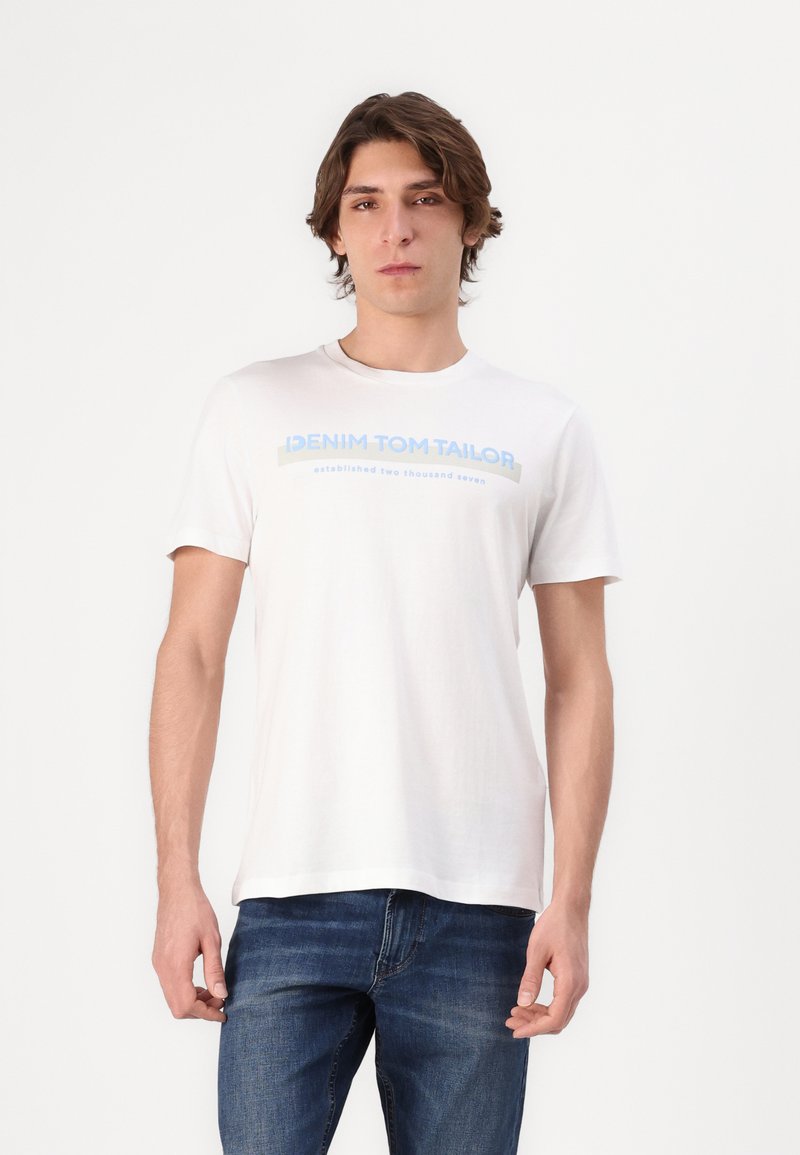 Valkoinen puuvillapaita lyhyillä hihoilla, jossa on sininen teksti "DENIM TOM TAILOR" ja "perustettu kaksituhattaseitsemän" edessä.