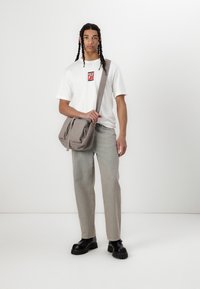 Grå messenger taske med to frontlomme med lynlås, i canvasmateriale, båret af en person i en hvid t-shirt og lyse jeans, med sort sko.