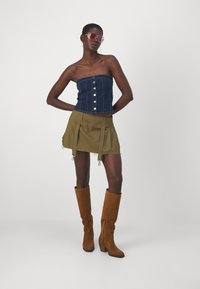 Denim strapless top with front buttons, paired with an olive green mini skirt with side zippers, and brown knee-high suede wedge boots.