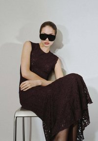 Abito in pizzo bordeaux scuro con collo alto e orlo asimmetrico. Modella indossa occhiali da sole neri oversize, seduta su una sedia di metallo.