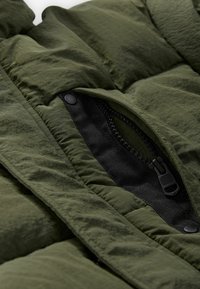 Grön puffjacka med detaljer som en dragkedjeficka med svart hårdvara, texturerad tyg med synlig sömnad och veck.