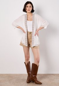 White sheer kimono with brown embroidered trim, paired with a white tank top and beige shorts. Brown cowboy boots complete the look.