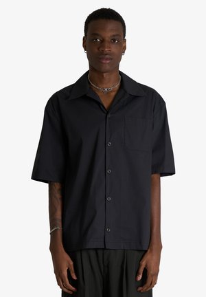 Giovane uomo con camicia nera a maniche corte abbottonata, collana a catena d'argento e orecchini, con tatuaggio sull'avambraccio sinistro, in piedi su sfondo bianco.