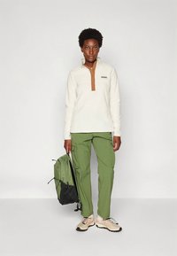 Fleece trui in beige met een bruine kraag, gecombineerd met olijfgroene cargobroek en beige wandelschoenen. Rugzak in groen en zwart.