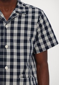 Κοντομάνικο πουκάμισο σε ναυτικό μπλε και λευκή καρόλα, με κομμένο γιακά, κουμπιά μπροστά και μία τσέπη στο στήθος.