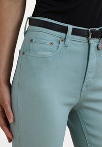 Vaqueros de mezclilla azul claro con un diseño ajustado, que cuentan con un cinturón negro, herrajes en tono dorado y bolsillos frontales con detalles de costura visibles.