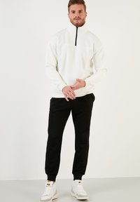 Fehér, magas galléros fleece pulóver fél cipzárral, fekete jogger nadrággal és fehér sportcipővel párosítva. Egyszerű, modern sportos öltözet.
