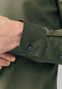 Camisa verde oliva con un primer plano del puño, que muestra botones azul oscuro y una tela suave y texturizada con detalles de costura precisos.