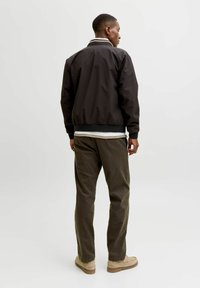 Άνδρας με μαύρο μπουφάν, ριγέ πουκάμισο, σκουρόχρωμο παντελόνι και μπεζ παπούτσια, με την πλάτη γυρισμένη, στέκεται μπροστά σε απλό φωτεινό φόντο.