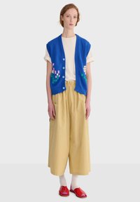 Mujer con chaleco de punto azul abotonado con diseño floral pixelado, camiseta blanca, pantalones anchos beige, calcetines blancos y zapatos rojos abiertos en los dedos, de pie.