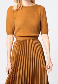 Kvinna iklädd en senapsgul stickad kortärmad tröja instoppad i en matchande plisserad midikjol mot en neutral bakgrund.