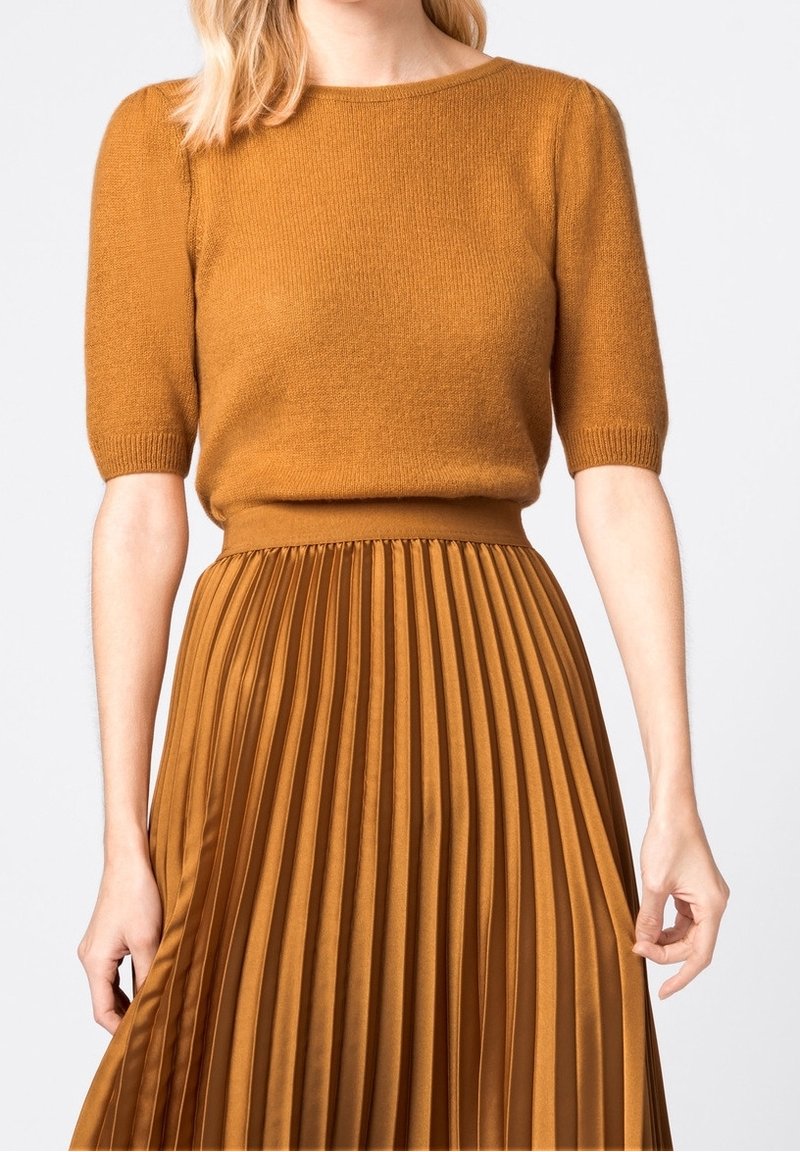 Kvinna iklädd en senapsgul stickad kortärmad tröja instoppad i en matchande plisserad midikjol mot en neutral bakgrund.