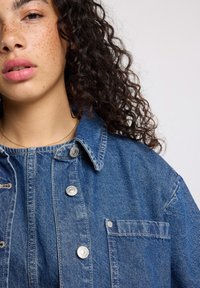 Giacca di denim di blu medio con chiusura a bottoni, colletto a punta, tasca sul petto e tessuto strutturato. Presenta bottoni in tonalità argento.