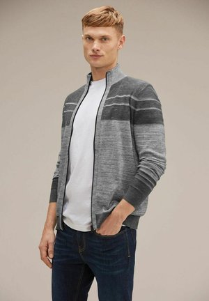 Cárdigan gris con cremallera y rayas horizontales, puños y cuello de canalé. Lleva puesto sobre una camiseta blanca, combinado con jeans oscuros. Tejido suave y texturizado.