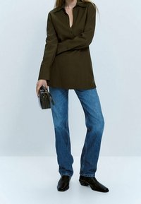 Chemise boutonnée vert olive avec un col, associée à un jean en denim bleu et des bottines noires, tenant un petit sac à main noir.