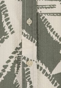 Camisa de botones de estampado verde y beige que presenta un tejido texturizado, costuras visibles y un botón de color crema.