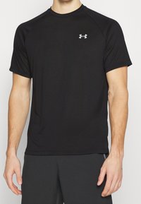 Svart kortärmad sporttröja tillverkad av mjukt, lätt tyg med rund hals och en liten grå logotyp på vänster bröst.