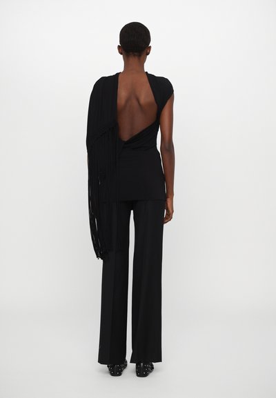 Top negro sin espalda con un diseño drapeado y detalle de flecos en un hombro, combinado con pantalones anchos negros y zapatos negros con tachuelas.