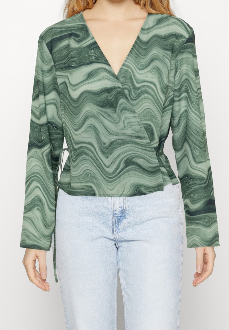 Top enroulé marbré vert avec des manches longues, présentant un décolleté en V profond et un détail de nouage sur le côté. Associé à un jean en denim bleu clair.