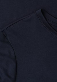 Mørk marineblåt stof, der viser en nærbillede af en T-shirts skuldersøm og rund halsudskæringens syning.