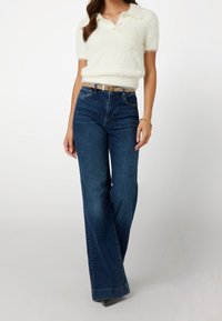 Biała, puszysta koszulka polo z krótkim rękawem, ciemnoniebieskie, biodrówki z rozszerzanymi nogawkami, zestawione z paskiem i szpiczastymi butami.