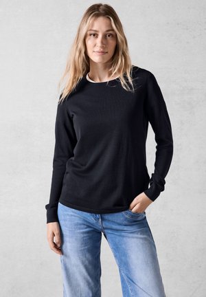 Chemise noire à manches longues avec col rond et poignets côtelés, associée à un jean en denim bleu clair, sur fond neutre.