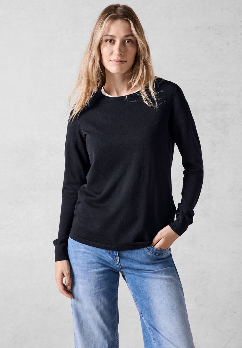Chemise noire à manches longues avec col rond et poignets côtelés, associée à un jean en denim bleu clair, sur fond neutre.
