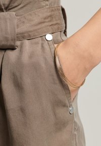 Bruine stof met een gladde textuur heeft een zijzak die is gesloten met een zilveren drukknoop. Een gouden armband rust op de pols.