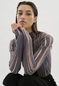 Vrouw met rechtgetrokken haar die een lange mouwen, semi-doorzichtige gestreepte top in paars en neutrale tinten draagt, met haar handen gekruist bij haar kin.