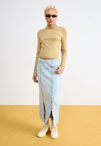 Lysebrun langærmet top med høj hals; lyseblå denimnederdel med lynlås foran og sidelommer; beige sneakers; gul gulv.