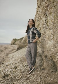 Sweter robiony na drutach w granatowo-kremową kratę, beżowe luźne spodnie i brązowe trampki. Modelka stoi na skalistej plaży.