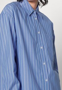 Hellblaues Hemd zum Knöpfen mit weißen vertikalen Streifen, einem Kragen und einer Brusttasche. Der Stoff wirkt glatt und leicht glänzend.