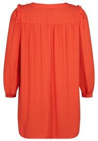 Tunique à manches longues de couleur orange uni, confectionnée dans un tissu léger. Elle présente un détail froncé aux épaules et des poignets boutonnés.