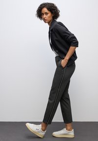 Ciemnoszare spodnie z białymi paskami po bokach, w połączeniu z czarną marynarką. Modelka w białych sneakersach, stojąca na jednolitym tle.