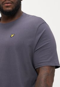 Graues T-Shirt mit kurzen Ärmeln aus Baumwolle und rundem Ausschnitt. Mit kleinem gelbem gesticktem Vogel-Logo auf der linken Brust.