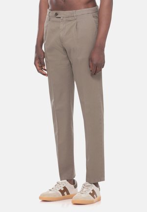 Uomo che indossa pantaloni beige a gamba dritta e scarpe da ginnastica bianche con dettagli marroni, in piedi davanti a uno sfondo neutro.