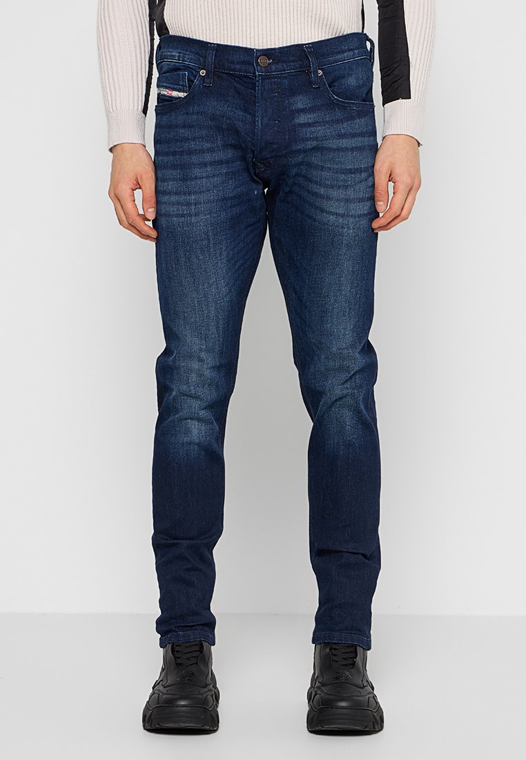 Jeans en denim bleu foncé avec une coupe ajustée, couture visible, poches avant et décoloration subtile. Associés à des chaussures noires.