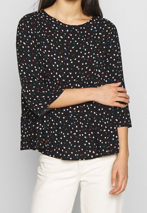 Femme portant un chemisier noir à manches longues avec des pois multicolores et un pantalon blanc, les bras croisés sur le torse.