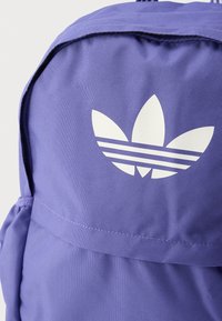 Μωβ σακίδιο πλάτης με υφή υφάσματος, που διαθέτει ένα εμφανές λευκό λογότυπο Adidas και τρεις οριζόντιες ρίγες στο κάτω μέρος.